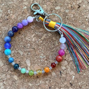 May include: A circular keychain featuring a strand of multicolored beads in a spectrum of shades, including purple, blue, green, yellow, orange, and red. The beads are attached to a silver-toned metal lobster clasp and a large split ring with a tassel.