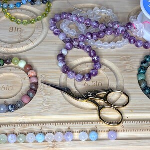 Puede incluir: Pulseras de cuentas de varios colores, incluyendo verde, morado y transparente, exhibidas sobre una superficie de madera con guías de medición. También hay tijeras doradas y cuentas sueltas.