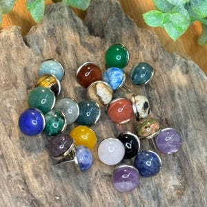 Op de afbeelding: Een verzameling ronde, gepolijste edelsteen punaises in diverse kleuren, waaronder blauw, groen, geel, oranje, paars, zwart, wit en bruin. Elke steen is gezet in een zilverkleurige metalen basis met een scherpe metalen pin aan de achterzijde.