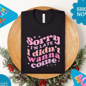 May include: Black t-shirt with the text "Sorry I'm late I didn't wanna come" in pink and orange retro font. The shirt is displayed on a wooden plate with a festive wreath. The shirt has a crew neck.
