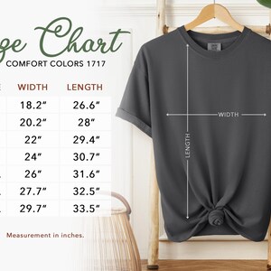 Puede incluir: Una camiseta de manga corta de color gris oscuro colgada en una percha de madera. A la izquierda hay una tabla de tallas que enumera de la S a la 4XL con medidas de ancho y largo en pulgadas, que van de 18,2 a 33,5 pulgadas.