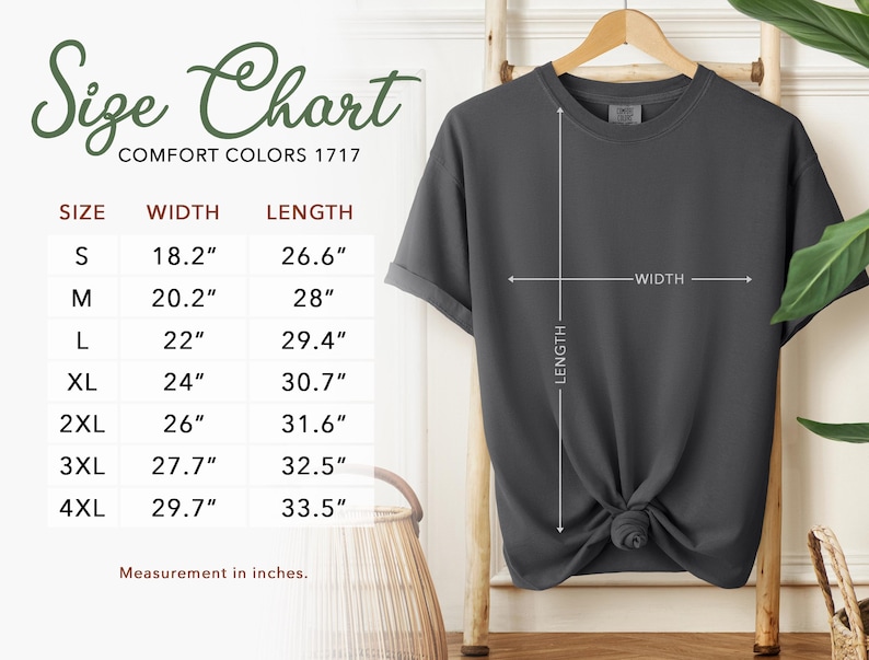 Puede incluir: Una camiseta de manga corta de color gris oscuro colgada en una percha de madera. A la izquierda hay una tabla de tallas que enumera de la S a la 4XL con medidas de ancho y largo en pulgadas, que van de 18,2 a 33,5 pulgadas.
