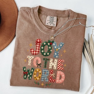 May include: A brown short-sleeve t-shirt featuring the embroidered text JOY TO THE WORLD in various patterned fabrics including red gingham, green floral, blue star, and plaid prints. The shirt has a Comfort Colors brand label at the neck.