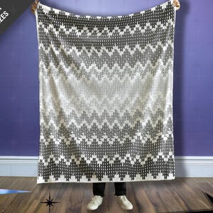 May include: A large, rectangular blanket with a geometric pattern in shades of black, gray, and white. The blanket is held up, displaying a chevron-like design. Text in the upper left corner reads "Choose from THREE SIZES."