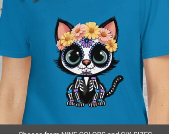 Könnte beinhalten: Ein türkisfarbenes T-Shirt mit einer Cartoon-Katze im Sugar-Skull-Design. Die Katze hat einen Blumenkranz und Skelett-Details. Der Text unten lautet "Choose from NINE COLORS and SIX SIZES."