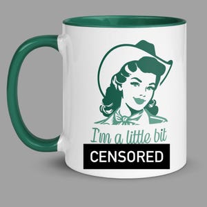 May include: White coffee mug with a green rim. The mug features a cartoon image of a woman wearing a cowboy hat and the text "I'm a little bit CENSORED".