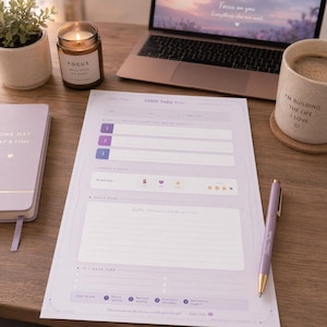 Op de afbeelding: Een bureau-opstelling met een lavendelkleurige ADHD Today Reset planner, een laptop, een kaars met "FOCUS" en een mok met "I'M BUILDING THE LIFE I LOVE". Een lavendelkleurige pen ligt op de planner. Een kleine potplant en een notitieboek zijn ook aanwezig.
