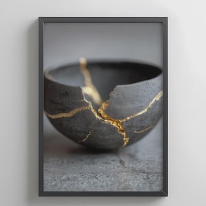 May include: A framed photograph of a black ceramic bowl, repaired with gold-coloured kintsugi. The bowl is cracked, with gold lines highlighting the fractures. The background is a soft grey, emphasising the bowl's form and texture.