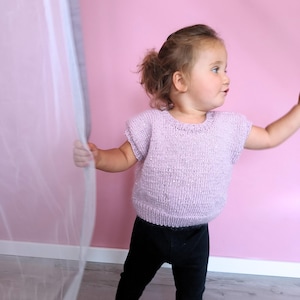 May include: A light lavender short-sleeved knit sweater with a round neckline and ribbed trim at the collar and hem, paired with solid black leggings. The sweater features a textured, speckled yarn finish and a simple, classic pullover construction.