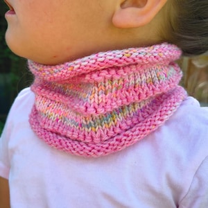 Könnte beinhalten: Ein rosa gestrickter Rundschal mit Rippenmuster. Der Schal ist aus weichem Garn gefertigt und weist eine Mischung aus Pastellfarben auf. Der Schal ist für das Tragen um den Hals konzipiert.