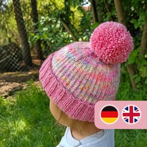 Può includere: Un cappello rosa lavorato a maglia con un grande pompon. Il cappello ha un bordo a coste e un motivo multicolore maculato. Il cappello è indossato da una persona, su uno sfondo di erba e alberi verdi.