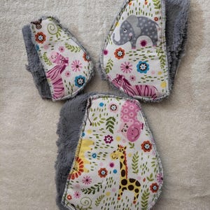 May include: Three fabric squares with a gray fuzzy backing. Each square features a different animal print with pink, yellow, and green flowers. The animals include a zebra, elephant, and giraffe.