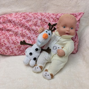 May include: A white baby doll with a light yellow onesie is holding a plush Olaf toy from the movie Frozen. The doll is lying on a pink floral pillow.