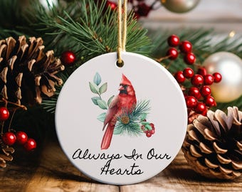 May include: A round white Christmas ornament with a red cardinal bird illustration, greenery, and the text "Always In Our Hearts." The ornament hangs from a gold string. The background includes pine needles, red berries, and pine cones.