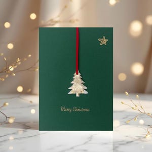 以下が含まれることがあります： 濃い緑色の背景のクリスマスカード。金色のクリスマスツリーのオーナメントが赤いリボンから吊り下げられています。金色の星が右上隅にあり、下部に「Merry Christmas」の文字があります。