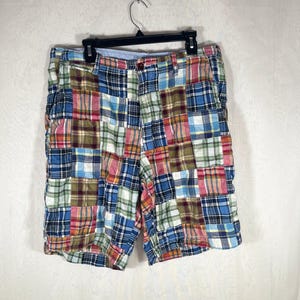 May include: Patchwork shorts featuring a multi-colored plaid pattern with squares of blue, red, green, yellow, and white fabric. The shorts have a button closure, a zipper fly, and belt loops, displayed on a black plastic hanger against a white background.