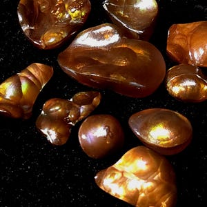 May include: A collection of polished fire agate gemstones featuring deep reddish-brown and amber hues with iridescent, glowing internal flashes of gold, orange, and green light. These smooth, organic-shaped stones exhibit a translucent quality.