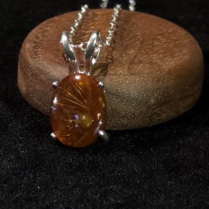 May include: An oval-shaped amber pendant featuring internal golden sun-like inclusions, set in a polished silver prong setting. The pendant hangs from a silver ball chain, showcasing a warm, translucent orange hue with distinct radiating patterns.