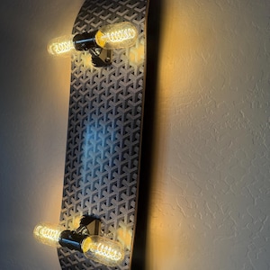 May include: A wall-mounted skateboard deck featuring a repeating black and grey geometric pattern. The board is equipped with two black truck mounts, each holding two elongated, glowing Edison-style light bulbs that emit a warm yellow light.