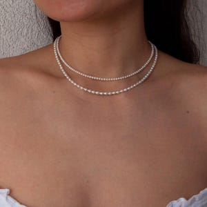 Op de afbeelding: Een dubbelstrengs ketting met twee lagen kleine, witte, ronde parels. De bovenste streng bestaat uit kleine, uniforme bolvormige parels, terwijl de onderste streng iets grotere, ovaalvormige parels bevat die tegen de hals rusten.