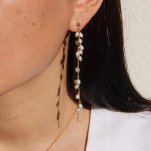 Op de afbeelding: Een goudkleurige oorbel met een waterval van kleine, witte parels. De oorbel hangt aan een kleine gouden bal, met de parels geregen langs een fijne ketting. Het ontwerp is elegant en minimalistisch.