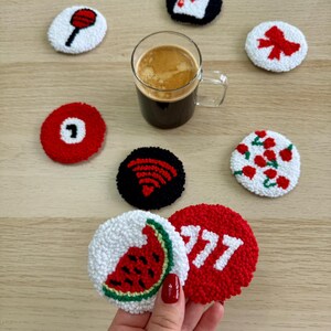 May include: A collection of handmade, round coasters with various designs, including a watermelon slice, a billiard ball, and playing card symbols. The coasters are white, red, and black, with a textured, tufted appearance. A glass of coffee sits among them.