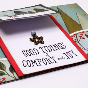 May include: A white card with the text "Good Tidings of Comfort and Joy" and a gold star ornament. The card is decorated with a red and green border and a background of blue and green trees.