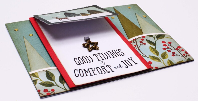 May include: A white card with the text "Good Tidings of Comfort and Joy" and a gold star ornament. The card is decorated with a red and green border and a background of blue and green trees.