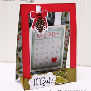May include: A red and green Christmas card with a pop-up calendar that says "Merry" and has the numbers 1-25. The card also has a pine cone with a red bow and the words "Peace on Earth" and "Joyful".