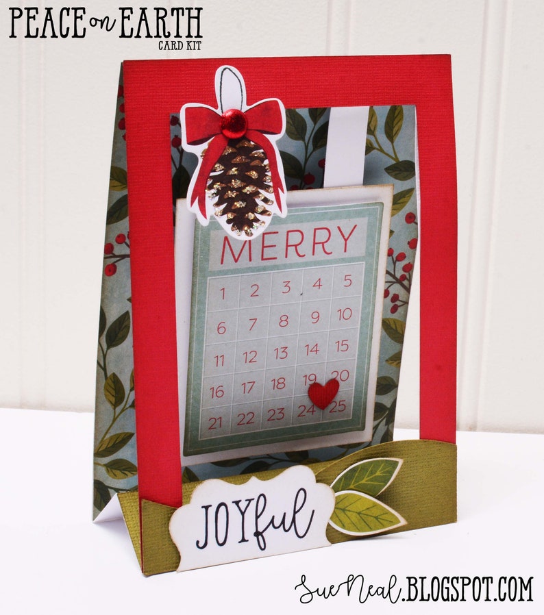 May include: A red and green Christmas card with a pop-up calendar that says "Merry" and has the numbers 1-25. The card also has a pine cone with a red bow and the words "Peace on Earth" and "Joyful".