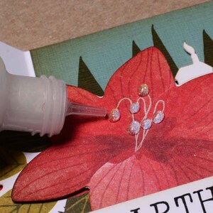 May include: A clear bottle of glitter glue with a silver cap and a long, thin nozzle is being used to add glitter to a red paper flower with white lines. The flower is part of a larger paper craft project.