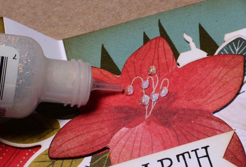 May include: A clear bottle of glitter glue with a silver cap and a long, thin nozzle is being used to add glitter to a red paper flower with white lines. The flower is part of a larger paper craft project.