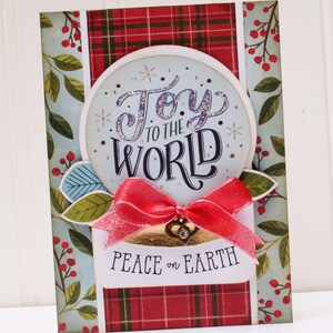 May include: A Christmas card with a red and green plaid background and a blue circle with the text "Joy to the World" in sparkly purple letters. The card also has a red ribbon and the text "Peace on Earth" in black letters.