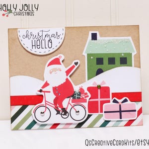 May include: A Christmas card with a red and white Santa Claus riding a bicycle with presents. The card has a green house with a white roof and the text "Christmas Hello" in a white circle.