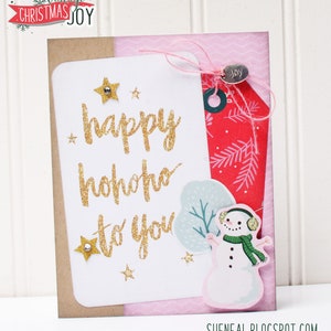 May include: A white card with gold glitter lettering that reads "happy hohoho to you". The card is decorated with gold glitter stars and a snowman sticker. The card is on a pink and brown background.
