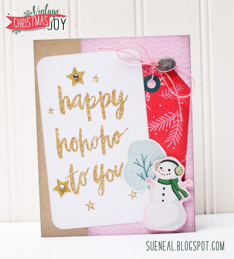 May include: A white card with gold glitter lettering that reads "happy hohoho to you". The card is decorated with gold glitter stars and a snowman sticker. The card is on a pink and brown background.