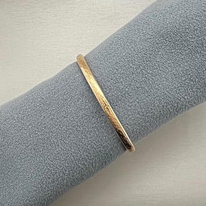 May include: A gold-colored bangle bracelet with a textured surface. The bracelet is resting on a gray fabric. The bangle is a simple, elegant piece of jewelry.