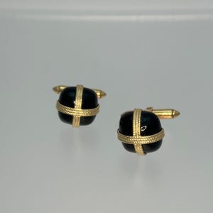 May include: A pair of cufflinks with a gold-colored metal base and a black, rounded center. The black center is encircled by a gold-colored band in a cross pattern. The cufflinks are displayed on a light-colored surface.