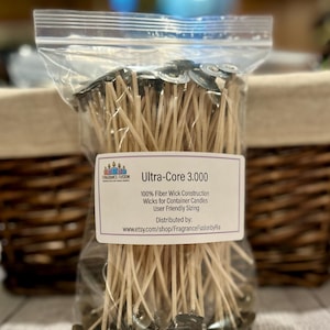 May include: A clear plastic bag filled with Ultra-Core 3.000 wicks for container candles. Each wick has a metal base and a beige fiber core. The label reads "100% Fiber Wick Construction" and "User Friendly Sizing".