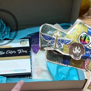 May include: An open box with blue tissue paper, containing a box of metallic color pens and a colorful, hand-drawn dragon head. The dragon head has various stickers and drawings, including a dragonfly and a cartoon character.