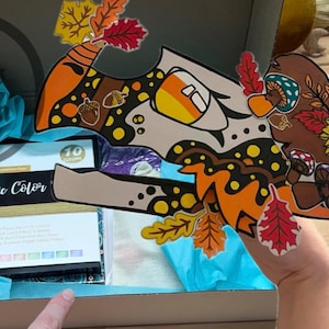 May include: An open box with a blue tissue paper lining. The box contains a set of 10 metallic paint markers and a colorful fall-themed paper craft. The paper craft features a brown bird with a sunflower, mushrooms, and leaves. The box also has the text "Metallic Color" printed on it.