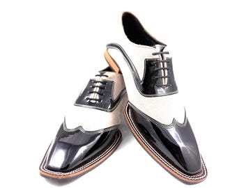 May include: A pair of two-tone oxford shoes with a unique design. The shoes feature a combination of black patent leather and off-white fabric, with black laces and a brown sole. The shoes have a distinctive, squared-off toe.