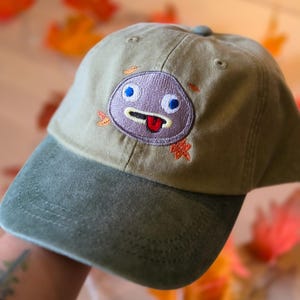 May include: An olive green baseball cap featuring an embroidered circular character with a grey face, blue eyes, and a red tongue sticking out. Small orange embroidered leaves surround the character on the front panel of the hat.