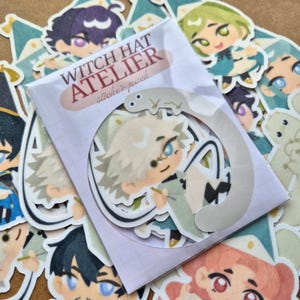 Puede incluir: Un paquete de pegatinas de la serie "Witch Hat Atelier". Las pegatinas presentan ilustraciones de estilo anime de personajes con varios colores de pelo y atuendos. El paquete de pegatinas está en una funda de plástico transparente con el nombre de la serie impreso.
