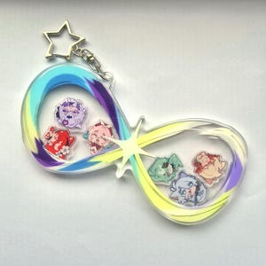 Könnte beinhalten: Ein klarer Acryl-Schlüsselanhänger in Form eines Unendlichkeitszeichens mit bunten Anime-Charms. Der Schlüsselanhänger hat einen sternförmigen Anhänger und einen Farbverlauf aus Blau, Gelb und Lila.