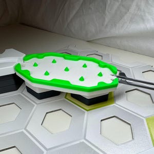 Könnte beinhalten: Eine weiße und grüne Spielbrettkomponente mit einem sechseckigen Muster. Die Komponente hat eine weiße Oberfläche mit grünen Akzenten und grünen konischen Formen. Die Komponente ist mit anderen grauen und weißen Spielteilen verbunden.