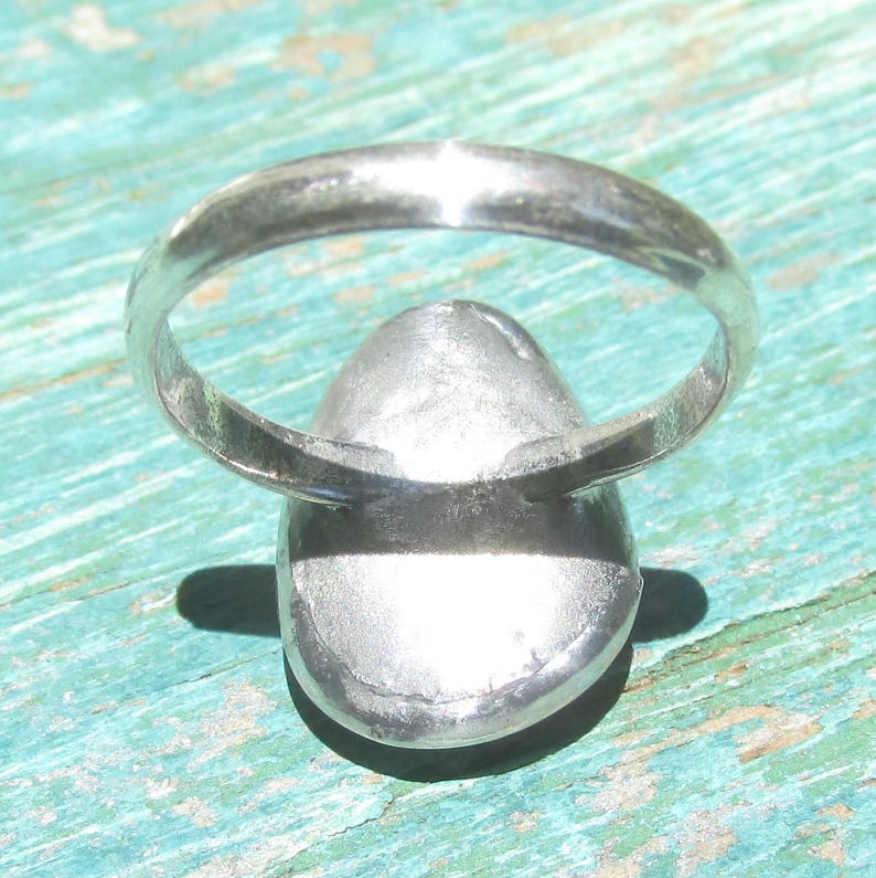 May include: A silver-toned ring featuring a smooth, rounded, metallic setting. The band is thin and circular, connecting to a solid, slightly domed, oval-shaped centerpiece. The metal surface has a reflective, polished finish throughout the entire piece.