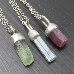 May include: Three gemstone pendant necklaces. One pendant is a light green crystal, the second is a blue crystal, and the third is a dark pink crystal. Each crystal is set in a silver-colored metal cap and hangs from a silver chain.