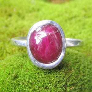 May include: A silver ring featuring a smooth, oval-shaped, deep magenta-red gemstone set in a polished metal bezel. The band is simple and metallic, showcasing a clean design that highlights the vibrant colour and reflective surface of the stone.