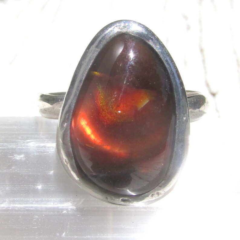 May include: A silver ring featuring a large, teardrop-shaped dark brown fire opal cabochon. The stone displays internal flashes of orange and red light. The metal setting is smooth and polished, encircling the gemstone in a simple bezel design.